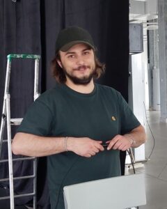 Un homme barbu à casquette devant un escabeau et derrière une chaise